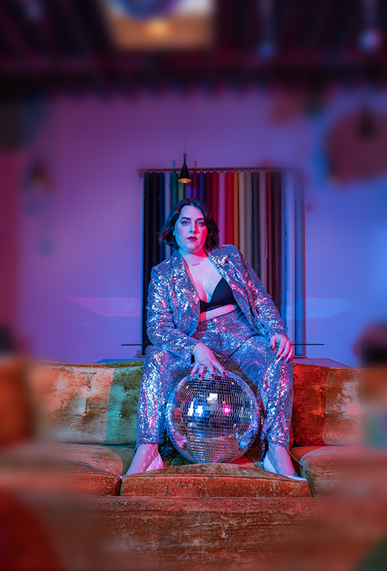 Picture of Hannah A. Patellis, a white woman with shoulder-length dark hair wearing a silver sequin pantsuit, sitting on the back of a retro couch with her hand on a large disco ball. The room is lit in reds, blues, and purples.