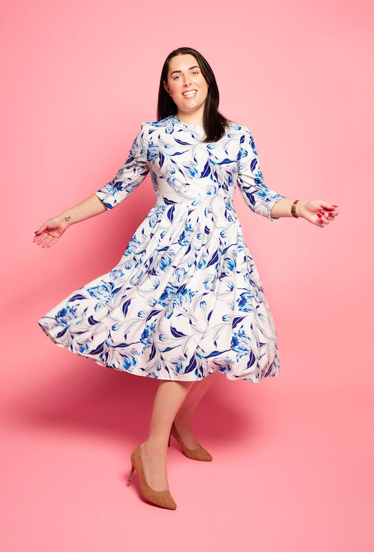 Picture of Hannah A. Patellis, a white woman with long dark hair, twirling in a white dress with blue flowers against a pink studio backdrop