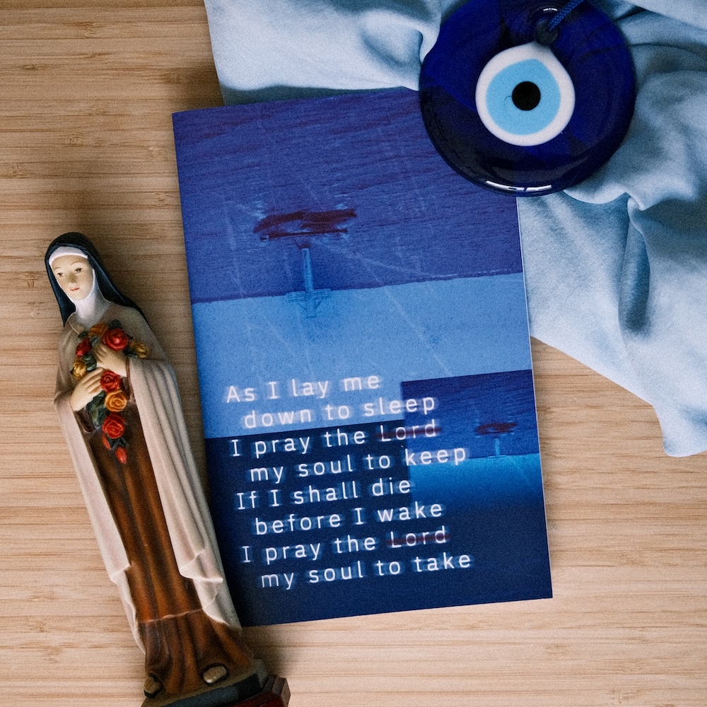 'As I Lay Me Down' zine cover featuring a distressed dark blue background with distressed upside-down photos up a light up cross on a lake and the words to the prayer 'As I lay me down to sleep.'