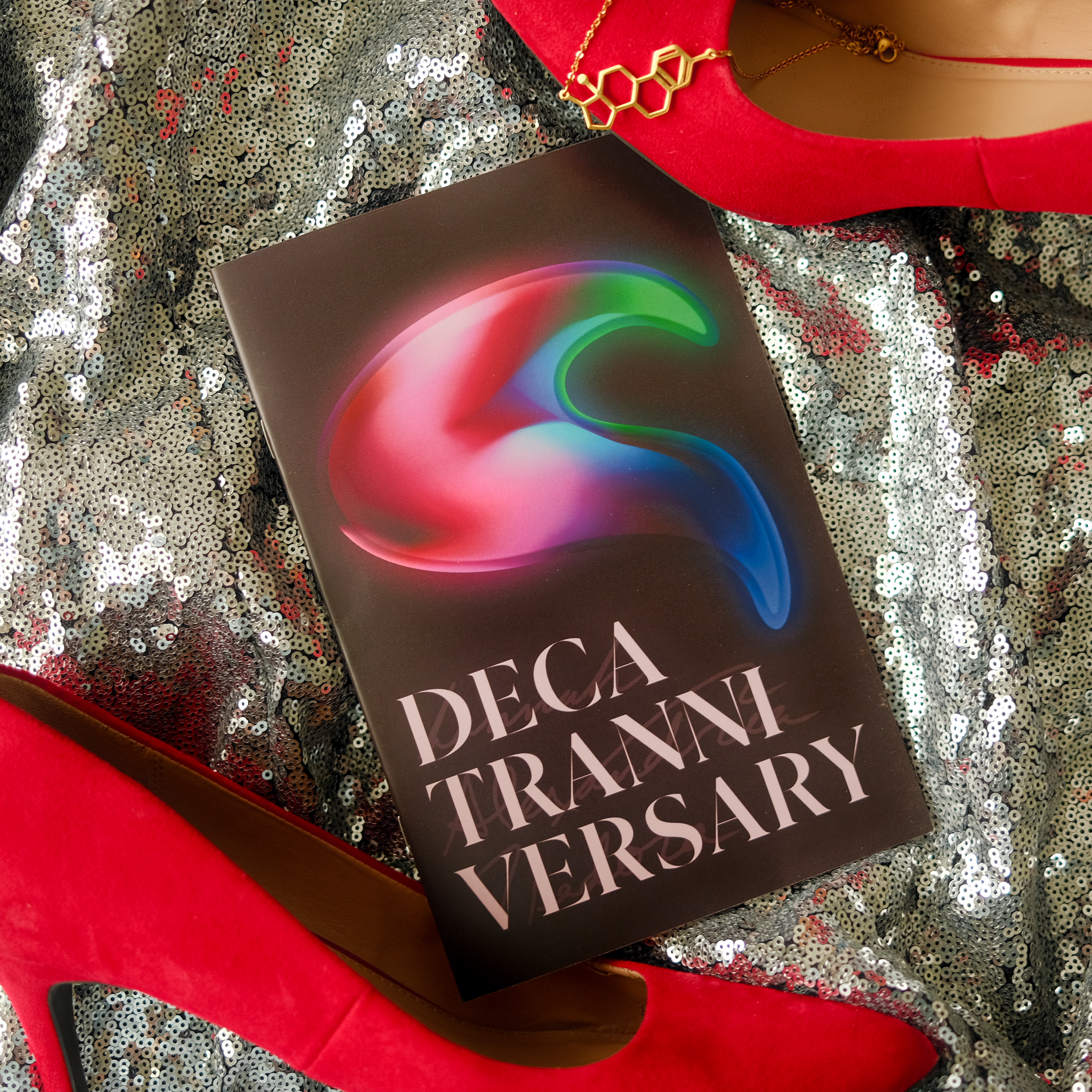 'DecaTranniversary' zine cover, a dark red background with a colorful abstract art piece taking up most of the page. The word 'DecaTranniversary' is on the bottom of the page in capital, pink, serif letters. Lightly behind the title is 'Hannah Alexandria Isadora' in cursive.